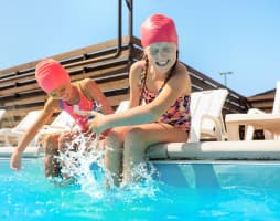 Two girls with swimming caps sitting on the edge of a pool, splashing their hands in the water and laughing.